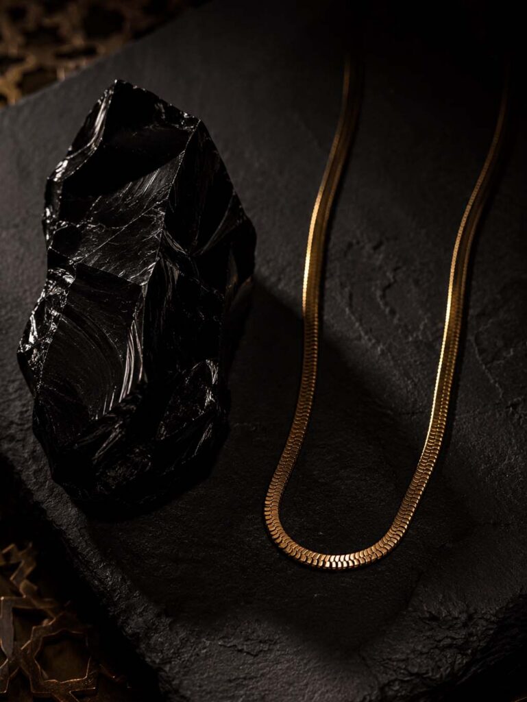Collier fine chaîne en or maille serpent posé sur une pierre noire texturée.